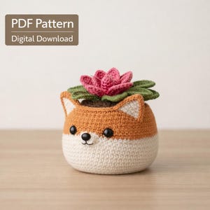 May include: A crocheted fox-shaped plant pot with a pink flower and green leaves. The pot is two-toned, with a cream base and orange top. The text "PDF Pattern Digital Download" is in the upper left corner.