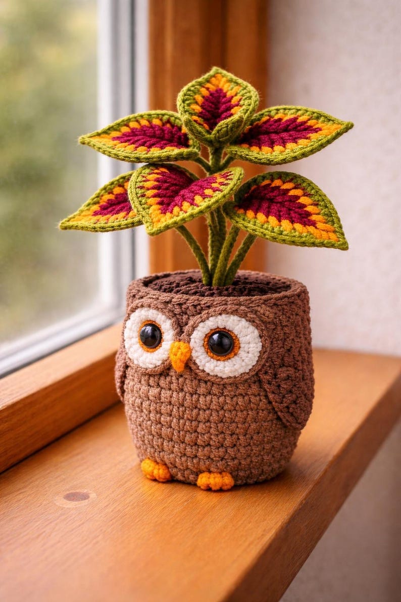 May include: A handcrafted crochet owl planter with a plant featuring green, yellow, and burgundy leaves. The owl planter is brown with large white-rimmed eyes, orange beak, and feet. The plant pot is sitting on a wooden surface.