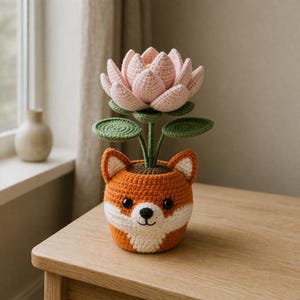 May include: A handmade crocheted fox plant pot in orange and white, with a pink lotus flower and green leaves. The fox pot has a smiling face and ears. The plant pot is on a wooden table.