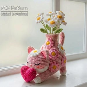 Op de afbeelding: Een roze gehaakte katvormige vaas met witte en gele madeliefjesaccenten, met een boeket witte en gele gehaakte bloemen. De kat houdt een roze hart vast. De tekst op de afbeelding luidt "PDF Pattern Digital Download".