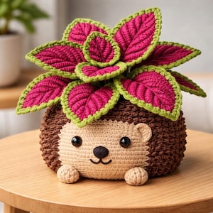 Może przedstawiać: Szydełkowy doniczka w kształcie jeża z rośliną o żywych różowych i zielonych liściach. Jeż ma beżową twarz, brązowe ciało i małe, okrągłe łapki. Liście rośliny mają szczegółowy, teksturowany wygląd.