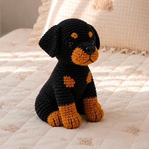 Pode incluir: Um peluche de croché feito à mão com a forma de um cachorro Rottweiler. O brinquedo é principalmente preto com marcas castanhas no focinho, acima dos olhos, no peito e na parte inferior das patas. Possui olhos e nariz de plástico preto.