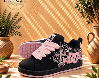 May include: A black DC sneaker with thick light pink laces, a pink DC logo on the side, and a dangling rhinestone cross charm. The shoe features a black suede-like upper with pink graphic patterns and a flat pink rubber sole.