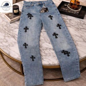 May include: Light blue denim jeans with multiple black cross appliqu&eacute;s. The jeans have a classic five-pocket design and a straight leg fit. The jeans are displayed on a marble-topped table.