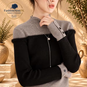 May include: A long-sleeved mock neck sweater featuring a color-blocked design with a grey upper section and a black lower section. The garment includes a fuzzy trim at the chest seam, grey cuffs with two small black buttons, and a silver-toned necklace.