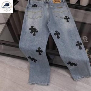 May include: Light blue denim jeans with black cross appliques. The jeans have a classic five-pocket design and a relaxed fit. The crosses are evenly spaced across the front and back of the jeans.