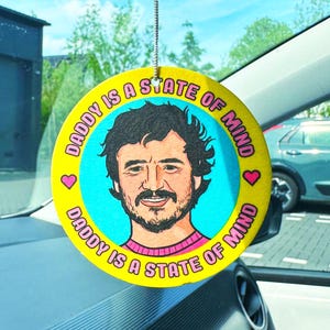 Puede incluir: Ambientador de coche redondo y amarillo con el texto "DADDY IS A STATE OF MIND" en un patrón circular. El centro presenta una ilustración de dibujos animados de un hombre con barba y bigote, con un suéter rosa. También incluye dos corazones rosas.