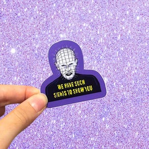 Peut inclure: Un autocollant violet découpé représentant une illustration en noir et blanc d'une tête pâle à motif quadrillé avec des épingles, portant une chemise noire. Le texte WE HAVE SUCH SIGHTS TO SHOW YOU est imprimé en lettres jaunes en bas.