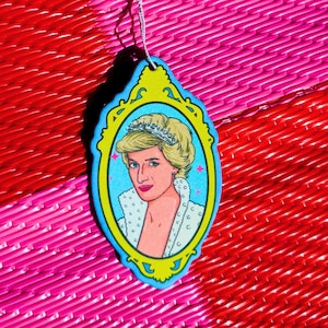 May include: A hanging ornament featuring a portrait of Princess Diana with blonde hair and a tiara, enclosed in a bright yellow and light blue ornate frame. The illustration shows a white collared garment with small dark dots and pink star accents.