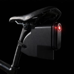 Puede incluir: Luz trasera de bicicleta negra montada en la tija del sillín. La luz roja está encendida. El sillín y la tija también son negros, sobre un fondo oscuro. Para mayor seguridad en bicicleta.