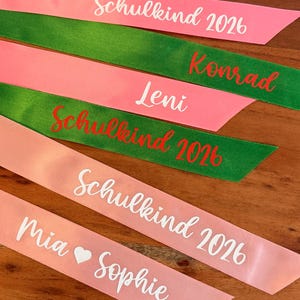 May include: Five satin ribbons in pink and green with custom text. The ribbons feature white or red lettering including the phrases Schulkind 2026, Konrad, Leni, and Mia Sophie. The ribbons are arranged in a parallel stack on a wooden surface.