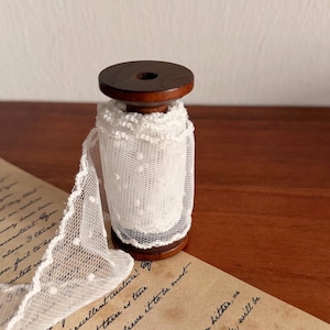 Könnte beinhalten: Eine Holzspule mit weißem Spitzenband. Die Spitze hat ein gepunktetes Muster und einen gewellten Rand. Die Spule ist dunkelbraun und liegt auf einem Stück gealtertem Papier mit handschriftlichem Text.