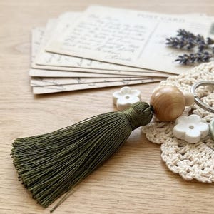 以下が含まれることがあります： 緑色のタッセル、木製のビーズ、葉のチャームが付いた手作りのキーホルダー。キーホルダーは、白い花の形をしたビーズが付いたかぎ針編みのドイリーの上にあります。ヴィンテージのポストカードとラベンダーの小枝が背景にあります。