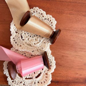 Puede incluir: Dos carretes de cinta de raso, uno beige y otro rosa, descansan sobre un tapete de ganchillo color crema. Los carretes tienen bases de madera oscura. La cinta está desenrollada, revelando su textura brillante. El fondo es una superficie de madera cálida.