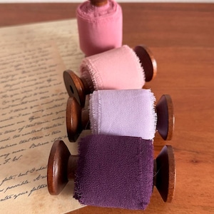 May include: Four spools of colorful ribbon in shades of pink, light pink, lavender, and purple. Each spool has a wooden base and top. The spools are arranged on a wooden surface, with a handwritten letter in the background.