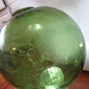 Pode incluir: Um grande flutuador de vidro verde, de forma esférica com a parte superior ligeiramente achatada. A superfície apresenta imperfeições e uma rede de finas rachaduras. Objeto decorativo, diâmetro de cerca de 25 cm, possivelmente náutico.