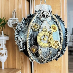 May include: Ornate steampunk-style Christmas ornament. The silver and black sphere is adorned with gold-colored gears, a pocket watch, and a rabbit figure. A silver metal cap and hanging chain complete the design. The ornament is approximately 4 inches in diameter.