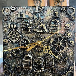 May include: A square wall clock with a textured black and gold finish, featuring raised numbers, various gear-shaped embellishments, and small sculpted doors. A central fairy figure sits above the clock hands, which are gold-colored metal.