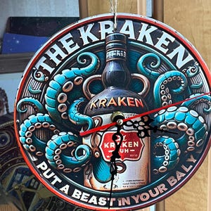 Op de afbeelding: Een ronde wandklok met een afbeelding van een Kraken rumfles omringd door blauwgroene tentakels. De tekst luidt THE KRAKEN en PUT A BEAST IN YOUR BELLY. De klok heeft zwarte uren- en minutenwijzers en een rode secondewijzer, opgehangen aan een touwtje.