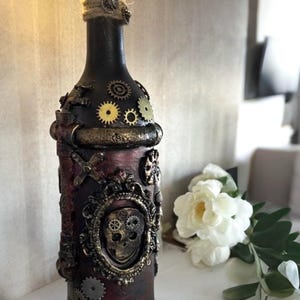 May include: A decorative steampunk-style bottle with a dark burgundy and black finish. The bottle is adorned with gears, a skull, and other metallic embellishments. The top is wrapped in twine and illuminated with fairy lights.