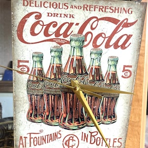 Op de afbeelding: Een rechthoekige wandklok met een vintage Coca-Cola-advertentie met vijf glazen flessen. De tekst luidt Delicious and Refreshing Drink Coca-Cola at Fountains in Bottles met twee 5-cent-symbolen. De klok heeft goudkleurige wijzers.