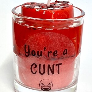 Op de afbeelding: Een rode kaars in een helder glas met de tekst "You're a CUNT" en een smiley. De kaars heeft een rode wasbasis en een rode bovenkant met glitter. Een witte lont steekt uit.