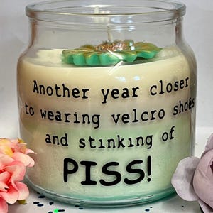 Pode incluir: Uma grande vela de vidro com um design de cera em camadas verdes e brancas. A vela tem uma flor decorativa verde e dourada no topo e o texto que diz "Another year closer to wearing velcro shoes and stinking of PISS!"