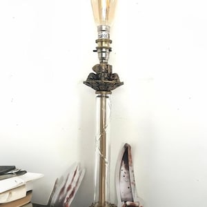 May include: A vintage table lamp with a decorative base and a clear glass column. The lamp features an Edison-style light bulb. Books and a decorative knife are in the foreground.