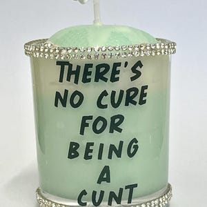 Op de afbeelding: Een mintgroene kaars met strass-details aan de boven- en onderkant. De kaars heeft de tekst "THERE'S NO CURE FOR BEING A CUNT" in zwarte letters. Bovenop de kaars zit een wit spiraalvormig ontwerp.