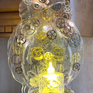 May include: A clear, owl-shaped decorative piece with embedded gears and a small, glowing candle. The owl is adorned with various sizes of metallic gears, creating a steampunk aesthetic. The candle sits on a decorative base, emitting a warm, yellow light.