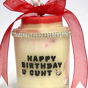 Op de afbeelding: Een kaars in een helder glas met een rode en witte bovenkant, een strassband en een rode strik. De kaars heeft de tekst "HAPPY BIRTHDAY U CUNT" in zwarte letters.