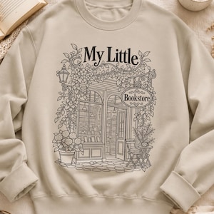 Könnte beinhalten: Ein beigefarbenes Sweatshirt mit Rundhalsausschnitt und einer schwarzen Strichzeichnung eines Ladenlokals mit dem Text "My Little Bookstore". Die Illustration zeigt einen Ladeneingang mit Kletterpflanzen, einem Topf und einem kleinen Tisch.