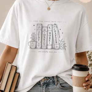 Könnte beinhalten: Ein weißes T-Shirt mit einer schwarzen Strichzeichnung von vier Büchern und einer Teetasse. Die Bücher sind mit dem Text "JUST ONE MORE CHAPTER" beschriftet und der Satz "FOR THE ONES WHO SAY ... and actually mean five." ist darüber und darunter gedruckt.