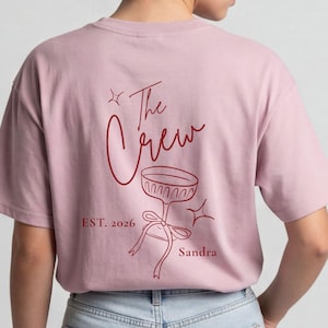 Op de afbeelding: Een lichtroze T-shirt met korte mouwen en een donkerrode afbeelding op de achterkant. Het ontwerp bevat de tekst "The Crew", "EST. 2026" en "Sandra" naast een lijntekening van een glas op een voet, versierd met een lint en sterren.