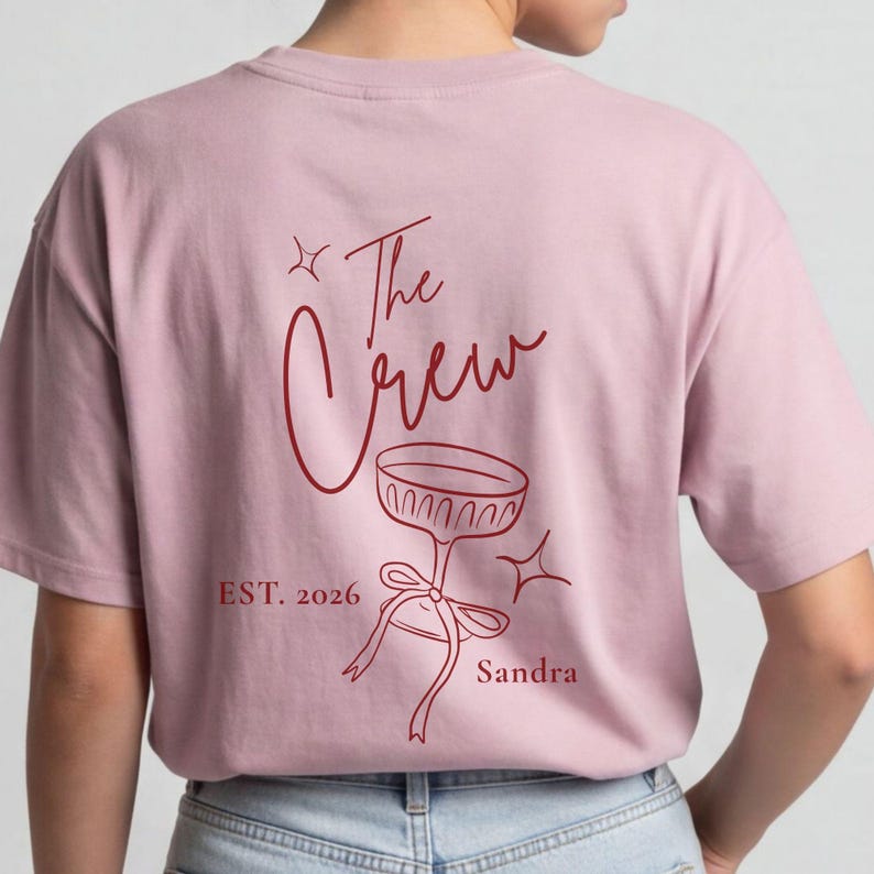 Op de afbeelding: Een lichtroze T-shirt met korte mouwen en een donkerrode afbeelding op de achterkant. Het ontwerp bevat de tekst "The Crew", "EST. 2026" en "Sandra" naast een lijntekening van een glas op een voet, versierd met een lint en sterren.