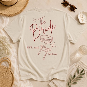 Op de afbeelding: Een cr&egrave;mekleurig T-shirt met korte mouwen en een rode afbeelding op de achterkant. Het ontwerp bevat de tekst The Bride, EST. 2026 en Melissa, naast een lijntekening van een glas op een voet met een strik en twee kleine sterren.