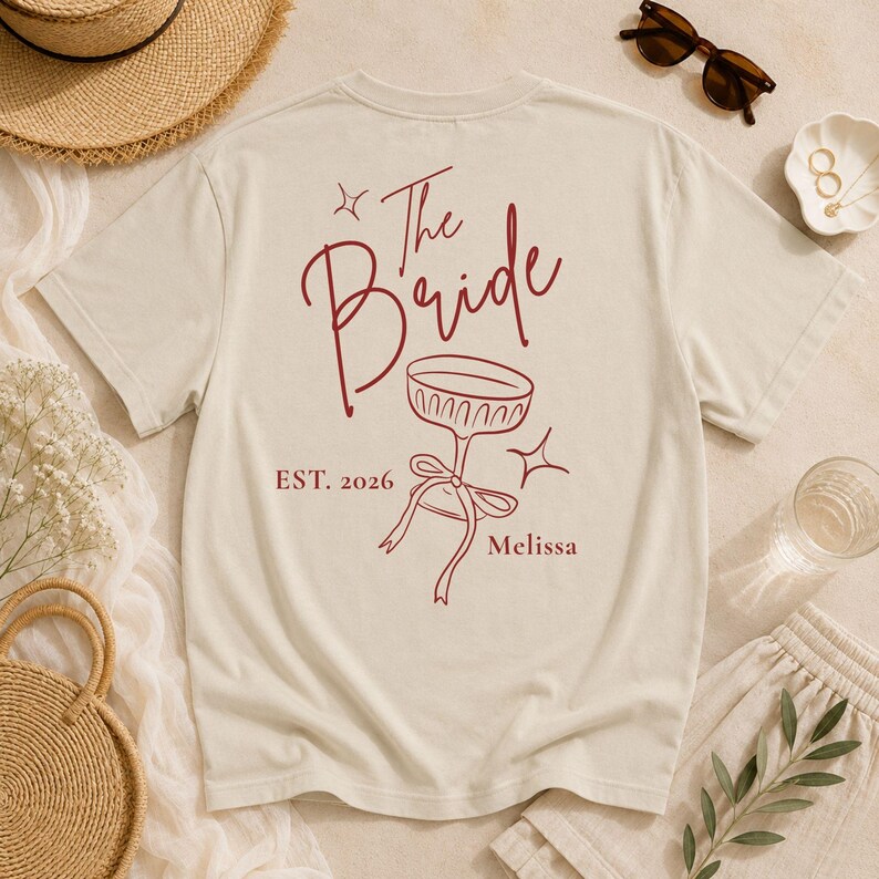 Op de afbeelding: Een cr&egrave;mekleurig T-shirt met korte mouwen en een rode afbeelding op de achterkant. Het ontwerp bevat de tekst The Bride, EST. 2026 en Melissa, naast een lijntekening van een glas op een voet met een strik en twee kleine sterren.