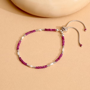 May include: A delicate bracelet featuring small, vibrant magenta beads and creamy white pearls. The bracelet is strung on a silver-toned chain with an adjustable closure. The design is simple and elegant.