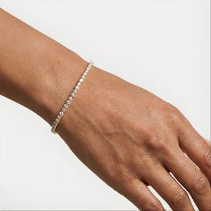 Puede incluir: Una pulsera de tenis dorada con numerosas piedras preciosas redondas y transparentes. La pulsera se lleva en la muñeca de una persona, con la mano y el brazo visibles sobre un fondo blanco. Es una joya delicada.
