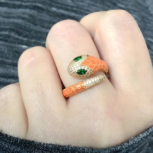 Puede incluir: Un anillo de serpiente en tono dorado con un diseño envolvente y escamas de esmalte color melocotón. La cabeza y el cuerpo están incrustados con pequeñas piedras de circonita cúbica transparentes y tiene dos piedras verdes talla marquesa.