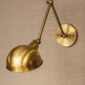 Puede incluir: Una lámpara de pared de latón con un brazo ajustable y una pantalla en forma de cúpula. La lámpara está fijada a una placa de pared redonda. El acabado es de un cálido color dorado metálico, y el diseño recuerda la iluminación industrial vintage.