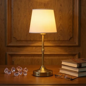 May include: A brass table lamp with a cream-colored lampshade. The lamp is on a wooden table with books, eyeglasses, and decorative crystals. The background is a wooden wall.