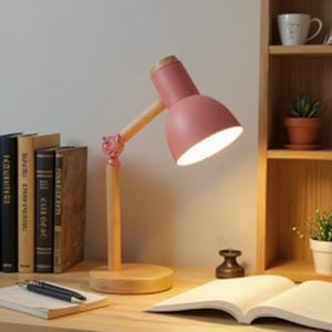 May include: A pink desk lamp with a wooden base and arm. The lamp has a round base, a wooden arm, and a pink lampshade. The lamp is turned on, illuminating the desk. The lamp is approximately 30 cm tall.