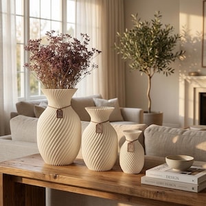 Könnte beinhalten: Drei cremefarbene Vasen unterschiedlicher Größe mit einem Spiraldesign, ausgestellt auf einem Holztisch. Die größte Vase enthält Trockenblumen. Jede Vase hat ein kleines Etikett. Wohnkultur.