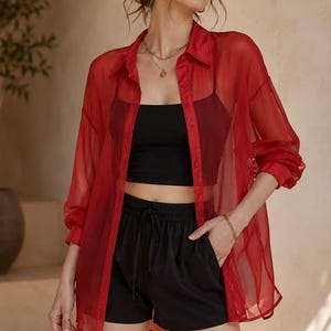 May include: A red sheer button-down shirt over a black crop top and black shorts. The shirt has long sleeves and is partially unbuttoned. The shorts have a drawstring waist. A gold necklace and bracelet are visible.