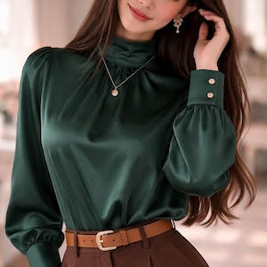 May include: A dark green satin blouse with a high neckline and long sleeves. The blouse has a slight sheen and is paired with brown pants and a brown belt. Gold buttons adorn the cuffs. A gold necklace is also visible.