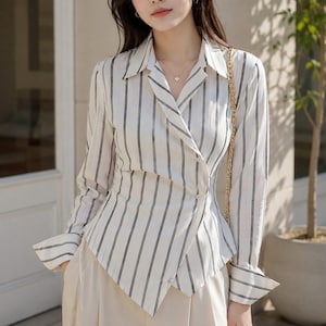 Puede incluir: Una camisa de manga larga blanca con finas rayas verticales negras. La camisa tiene un cuello en punta, un frente cruzado con un dobladillo diagonal y pu&ntilde;os anchos. Se combina con pantalones de color beige claro y una cadena dorada.