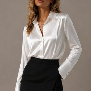 Puede incluir: Una camisa blanca de satén con mangas largas y una falda negra. La camisa tiene un cuello clásico y puños abotonados. La falda es negra y tiene un diseño cruzado con una abertura.