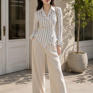 Puede incluir: Una camisa de manga larga con botones y un patr&oacute;n de rayas verticales blancas y negras, con cuello en punta y dobladillo asim&eacute;trico, combinada con pantalones de color crema de talle alto y pernera ancha y sandalias blancas abiertas.