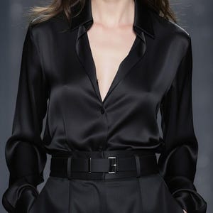 Pode incluir: Uma camisa preta de cetim com gola e mangas compridas, combinada com calças pretas e um cinto a condizer. A camisa está parcialmente desabotoada, criando um decote em V. O cinto tem uma fivela prateada.